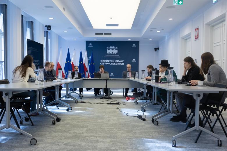 Kilkanaście osób siedzi przy stołach ustawionych w podkowę na sali konferencyjnej