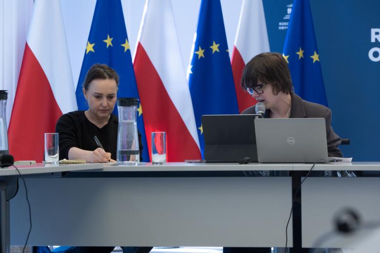Dwie osoby siedzą za stołem w sali konferencyjnej na tle flag polski i UE, jedna z nich mówi do trzymanego w dłoni mikrofonu
