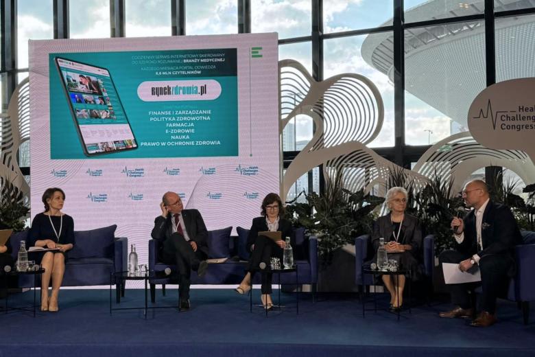 Kilka osób siedzi na fotelach na scenie w jasnej, przeszklonej sali konferencyjnej