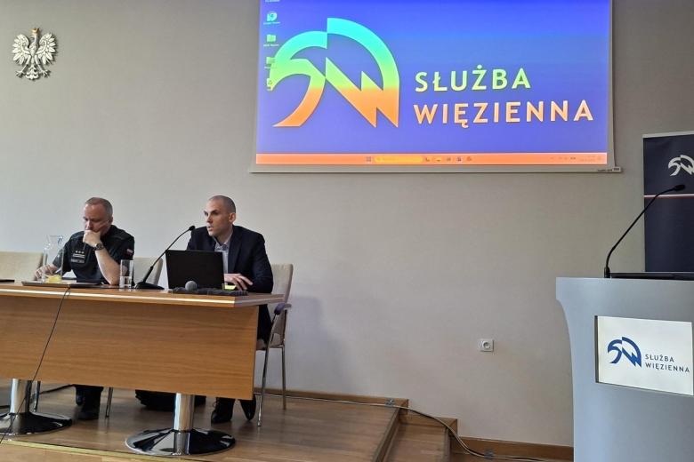 dwie osoby siedzą w sali za stołem, jedna przemawia 