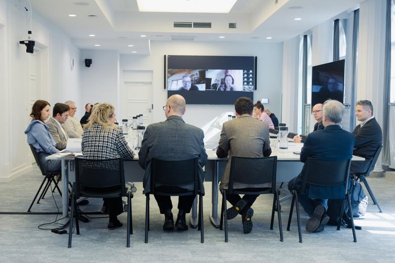 Kilkanaście osób siedzi przy stole w sali konferencyjnej. W szczycie stołu jest ekran, na którym wyświetlają się uczestnicy spotkania łączący się z pomocą internetu