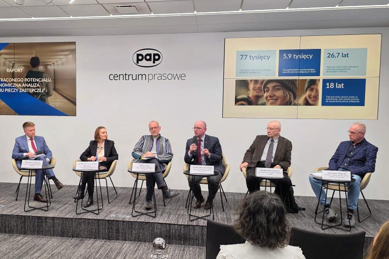 Sześć osób siedzi przez publicznością na krzesłach na podwyższeniu w salce konferencyjnej, jedna z nich mówi do trzymanego mikrofonu