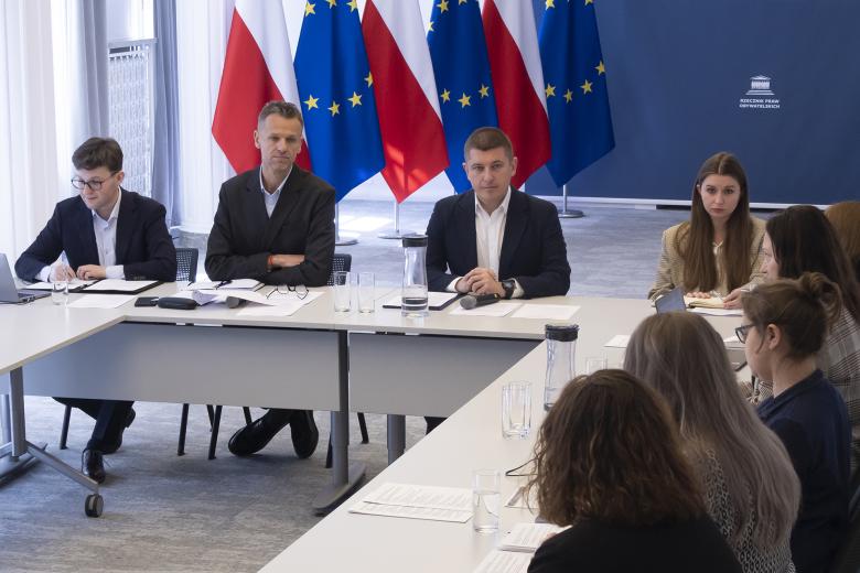 Kilka osób siedzi przy stołach ustawionych w podkowę na sali konferencyjnej i rozmawia, za stołem flagi Polski i UE