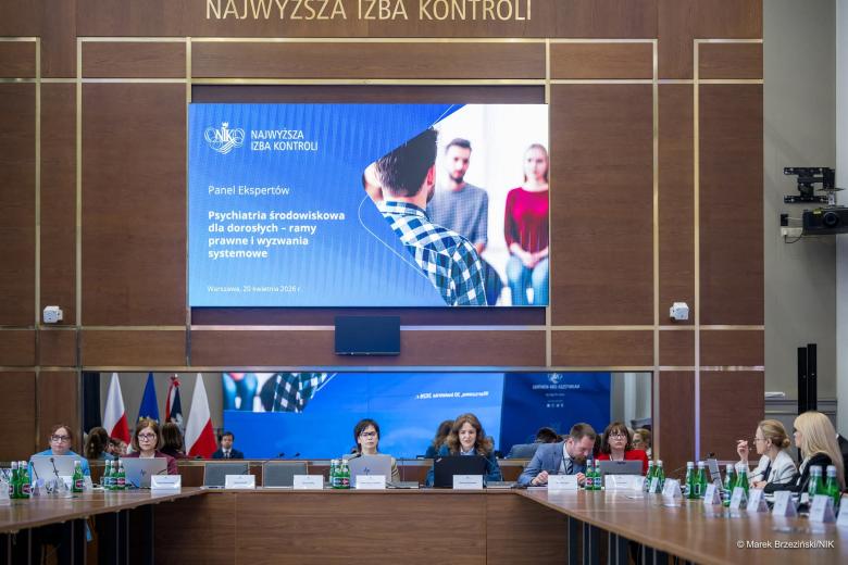 Dziesięć osób siedzi przy stole konferencyjnym. Nad stołem jest ekran, na którym wyświetla się nazwa spotkania: Psychiatria środowiskowa dla dorosłych - ramy prawne i wyzwania systemowe