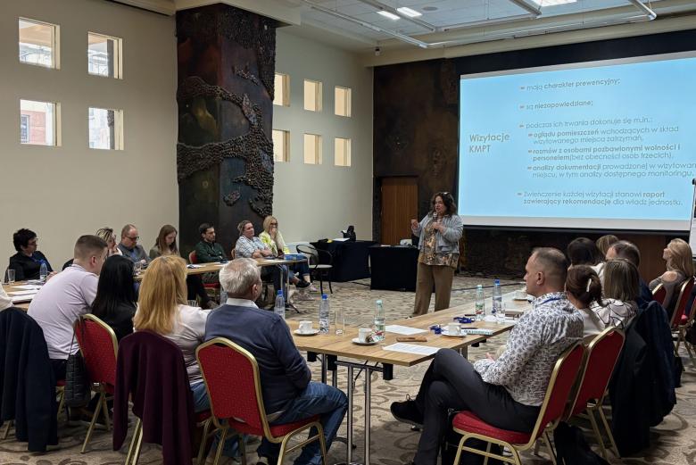 Kilkanaście osób siedzi przy stołach ustawionych w podkowę. Między stołami stoi kobieta z mikrofonem wygłaszająca prezentację, której treść wyświetla się na znajdującym się za nią dużym ekranie