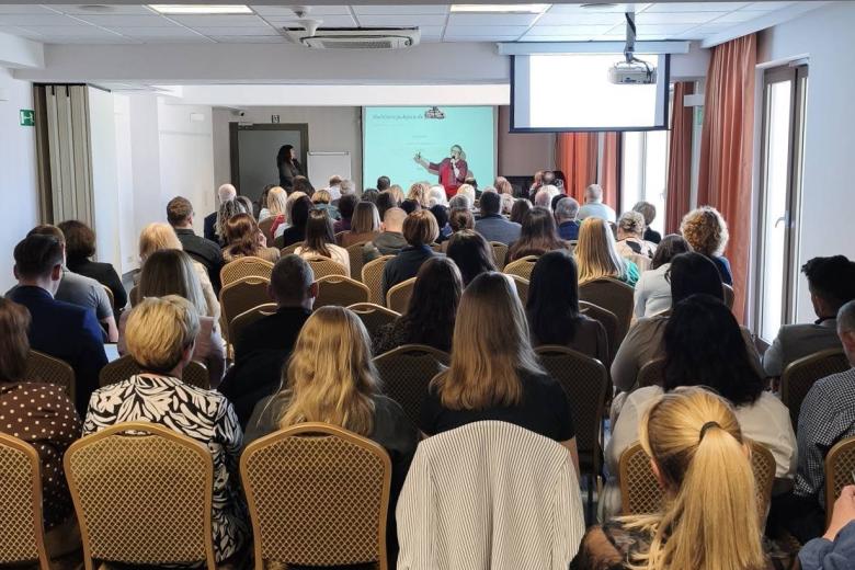 Kilkadziesiąt osób siedzi na krzesłach w sali konferencyjnej. W szczycie sali kobieta wygłasza prezentację na stojąco