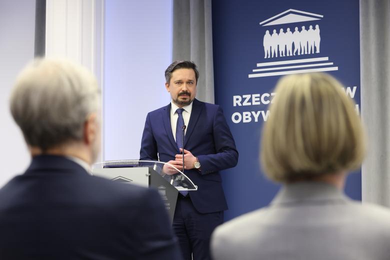 Marcin Wiącek stoi przy mównicy i wypowiada się w stronę siedzącego publiczności