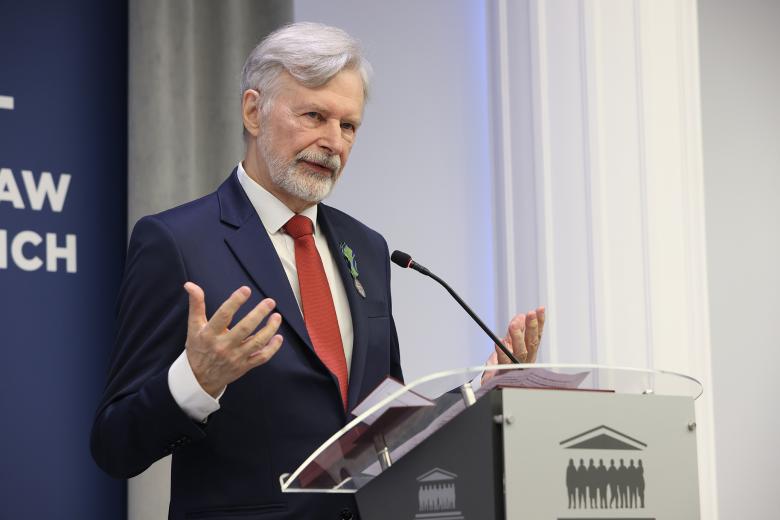 Marek Balicki wypowiada się stojąc przy mównicy. Na piersi ma przypięte odznaczenie.