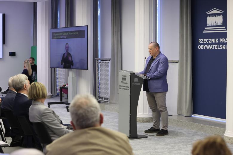 Łukasz Faliszek wypowiada się stojąc przy mównicy. Przed nim widoczne są osoby siedzące na widowni