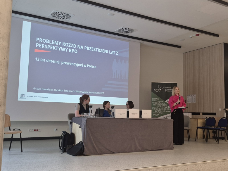 osoby siedzą w sali jedna przemawia na stojąco