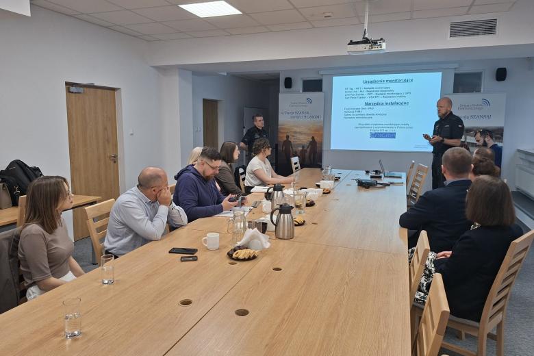 osoby siedzą w sali patrząc na ekran w tle