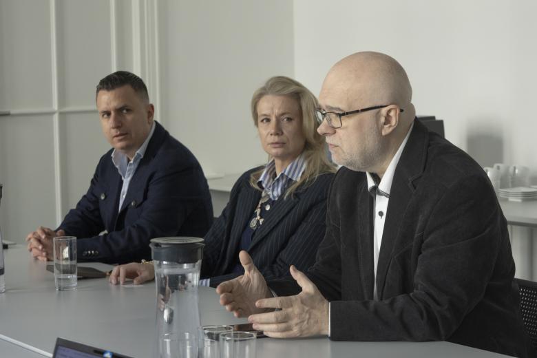 trzy osoby siedzą w sali jedna przemawia na siedząco 