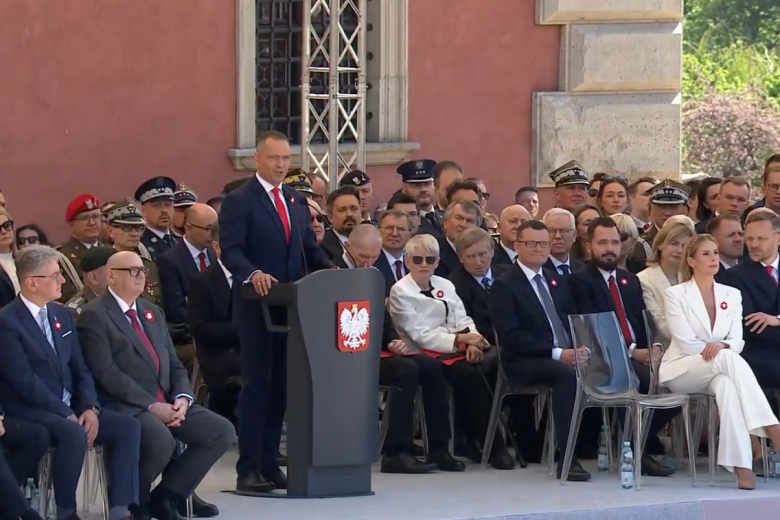 Na ustawionych na trybunie krzesłach siedzą uczestnicy obchodów. Przed nimi przy mównicy stoi prezydent Karol Nawrocki i wygłasza wystąpienie.