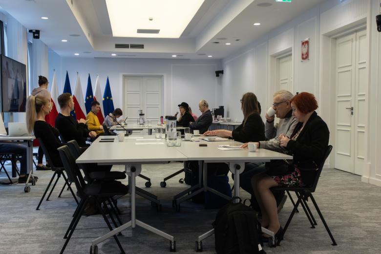 Kilkanaście osób siedzi przy stołach na dużej sali konferencyjnej