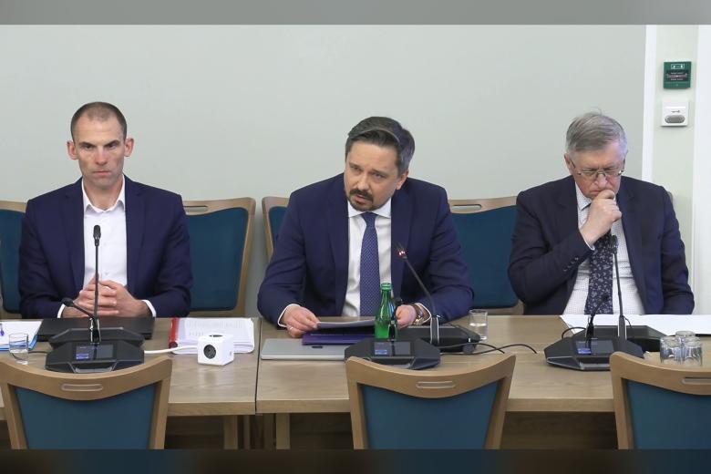 Trzy osoby siedzą za stołem, na którym leżą dokumenty i stoja mikrofony