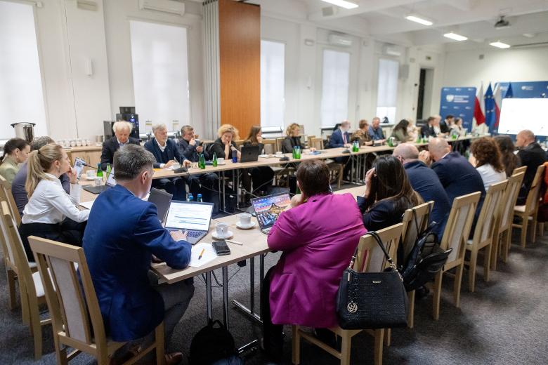 Kilkadziesiąt osób siedzi przy stołach ustawionych w podkowę na dużej sali konferencyjnej