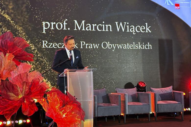 Osoba przemawia za mównicą na nastrojowo oświetlonym podium, w tle duży napis "prof. Marcin Wiącek, Rzecznik Praw Obywatelskich"