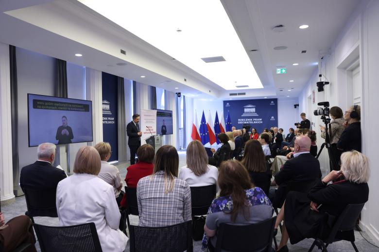 Kilkadziesiąt osób na sali konferencyjnej słucha wystąpienia osoby mówiącej za mównicą