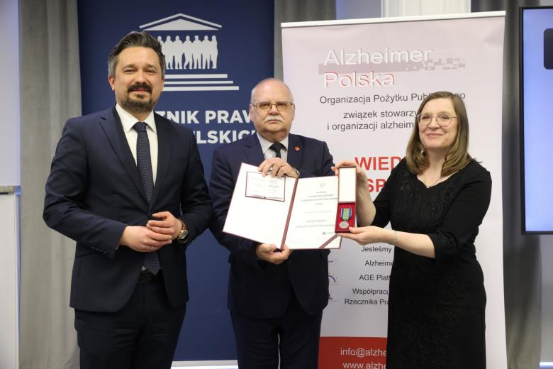 Trzy osoby pozują do wspólnego zdjęcia trzymając dyplom oraz odznakę w formie medalu