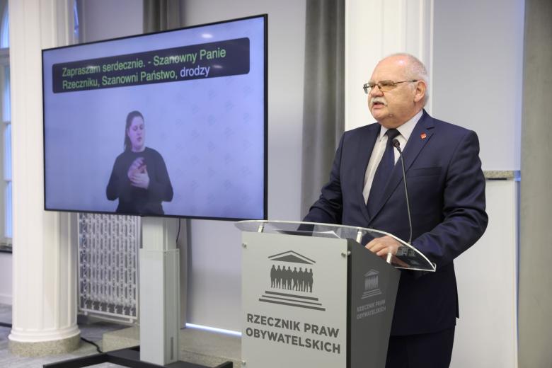 Osoba przemawia za mównicą z napisem "Rzecznik Praw Obywatelskich", w tle ekran z widocznym tłumaczem na Polski Język Migowy
