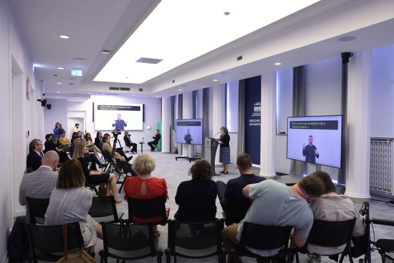 Kilkadziesiąt osób siedzi na sali konferencyjnej i słucha wystąpienia osoby mówiącej za mównicą na środku