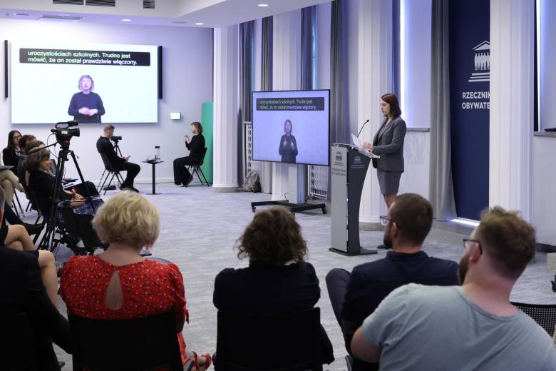 Osoba przemawia za mównicą na sali konferencyjnej, na której siedzi kilkadziesiąt osób