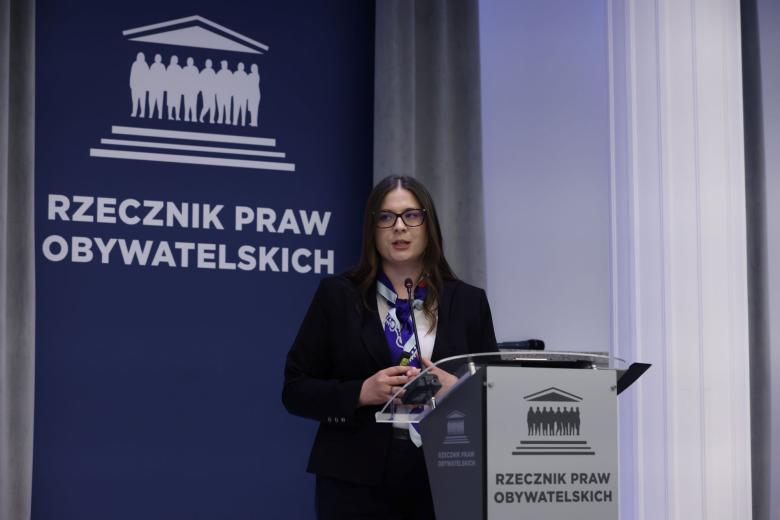 Osoba mówi za mównicą na sali konferencyjnej na tle planszy z napisem "Rzecznik Praw Obywatelskich"