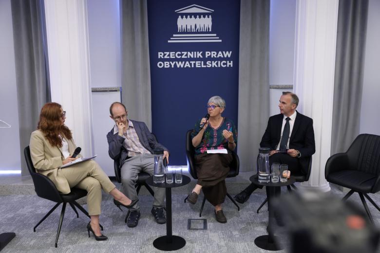 Cztery osoby siedzą na fotelach na sali konferencyjnej na tle planszy z napisem "Rzecznik Praw Obywatelskich", jedna z nich mówi do trzymanego w dłoni mikrofonu