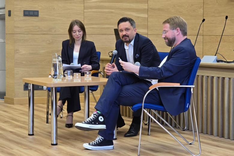 Trzy osoby siedzą przy stoliku na scenie auli wykładowej