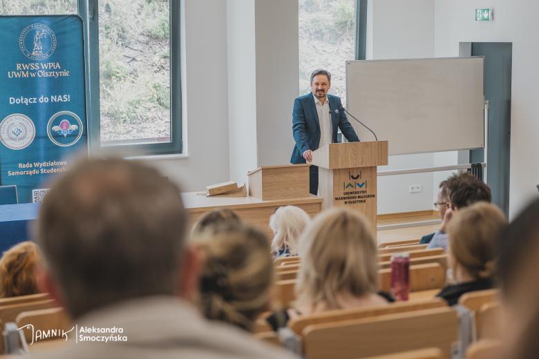 Osoba przemawia za mównicą na auli wykładowej. Na mównicy napis "Uniwersytet Warmińsko-Mazurski w Olsztynie"