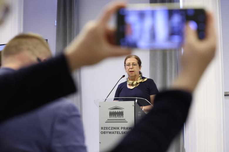 Małgorzata Czajkowska-Kisil wypowiada się stojąc przy podium. Na pierwszym planie widać dłonie osoby z publiczności trzymające telefon, którym robione jest zdjęcie 