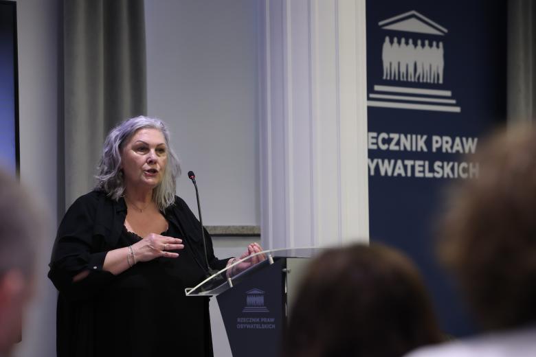 Elżbieta Dzik wypowiada się stojąc przy mównicy i gestykulując