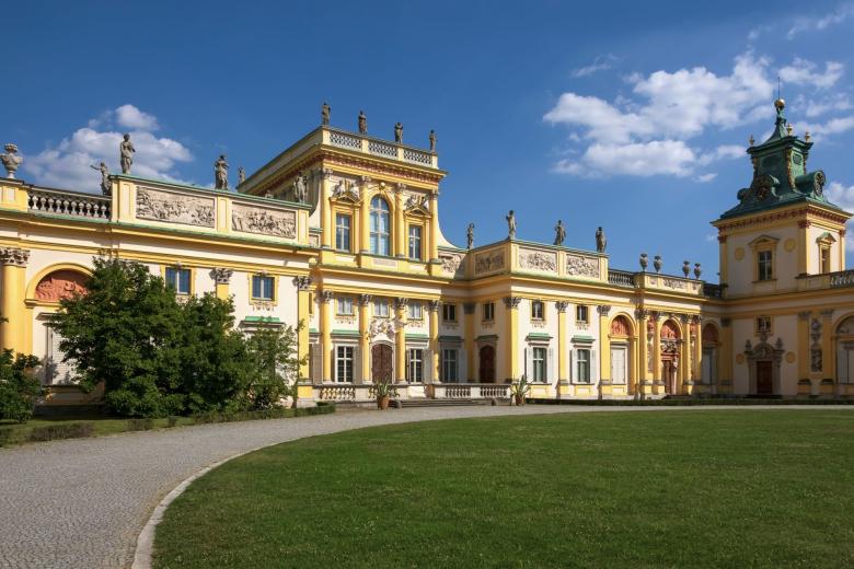 Zabytkowy, barokowy, zdobiony detalami architektonicznymi i rzeźbami budynek pomalowany na żółto i biało, z charakterystycznymi wieżami widoczny od strony dziedzińca z zadbanym trawnikiem