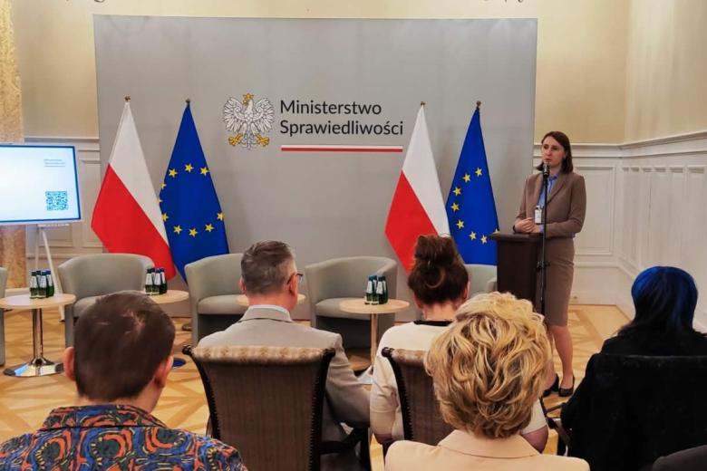 Osoba przemawia za mównicą na salce konferencyjnej, w tle flagi Polski i UE oraz duża plansza z godłem Polski i napisem "Ministerstwo Sprawiedliwości", wystąpienia słuchają osoby siedzące na krzesłach
