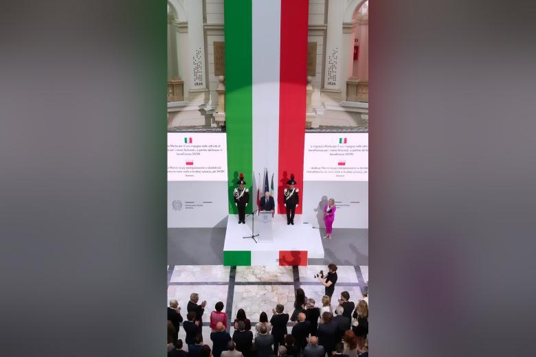 Zdjęcie z góry dużego, zabytkowego pomieszczenia z osobą przemawiającą za mównicą na podium na tle pionowo wiszącej flagi Włoch w kolorach zielonym, biąłym i czerwonym, wystąpieniu przysłuchują się zgromadzone w pomieszczeniu osoby