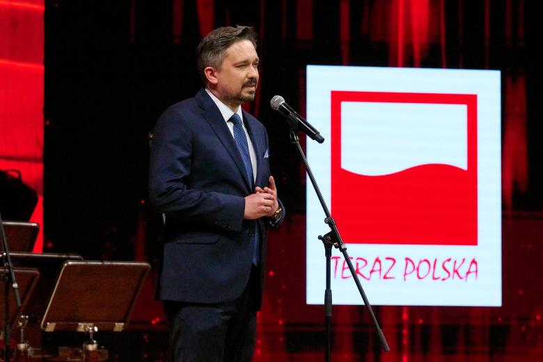 Osoba przemawia na stojąco na scenie, w tle znak graficzny z biało czerwoną flagą i napisem "Teraz Polska"