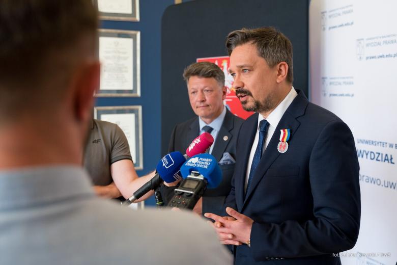 Osoba mówi do trzymanych przez dziennikarzy mikrofonów ze znakami graficznymi różnych stacji radiowych.