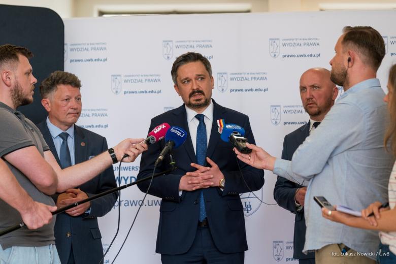 Kilka osób stoi na tle ścianki z logotypem Uniwersytetu w Białymstoku, jedna z nich mówi do trzymanych przez dziennikarzy mikrofonów