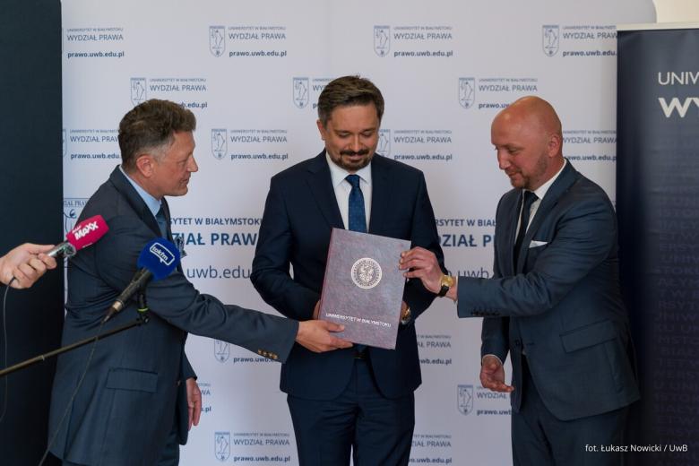 Trzy osoby stoją i pozują do zdjęcia na stojąco z teczką na dokumenty z napisem "Uniwersytet w Białymstoku"