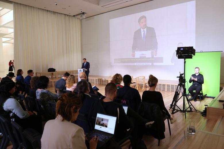 Widok sali konferencyjnej, w której siedzi kilkadziesiąt osób. Przed nimi przy mównicy stoi mężczyzna i wypowiada się, po prawej stronie na scenie na zielonym tle widoczny tłumacz PJM