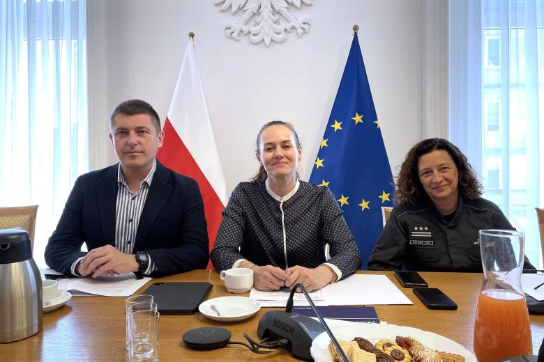trzy osoby siedzą w sali na tle flag Polski i UE 