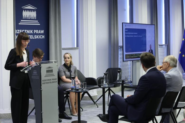 Osoba przemawia za mównicą z napisem "Rzecznik Praw Obywatelskich", obok niej na krzesłach siedzą dwie osoby, a z przodu widownia przysłuchująca się wystąpieniu
