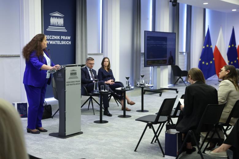 Osoba przemawia za mównicą z napisem "Rzecznik Praw Obywatelskich", obok niej na krzesłach siedzą dwie osoby, a z przodu widownia przysłuchująca się wystąpieniu