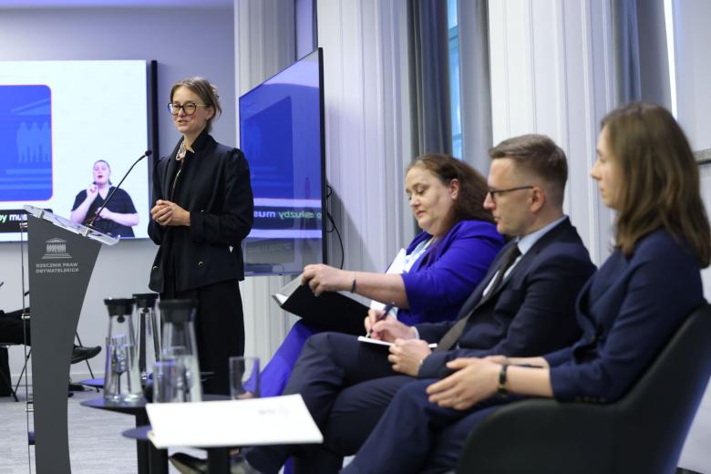 Osoba przemawia za mównicą z napisem "Rzecznik Praw Obywatelskich", obok niej na krzesłach siedzą trzy osoby