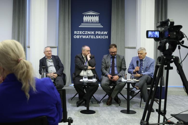 Cztery osoby siedzą na krzesłach przy małych stolikach, w tle granatowa plansza z napisem "Rzecznik Praw Obywatelskich"