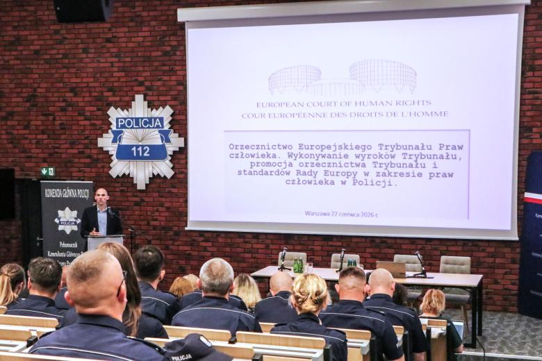 Kilkudziesięciu policjantów w mundurach słucha wystąpienia osoby mówiącej za mównicą na sali wykładowej