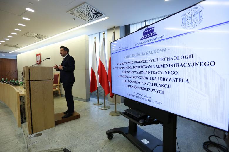 Osoba przemawia za mównicą, na pierwszym planie duży wyświetlacz z tytułem konferencji i logotypami organizatorów.