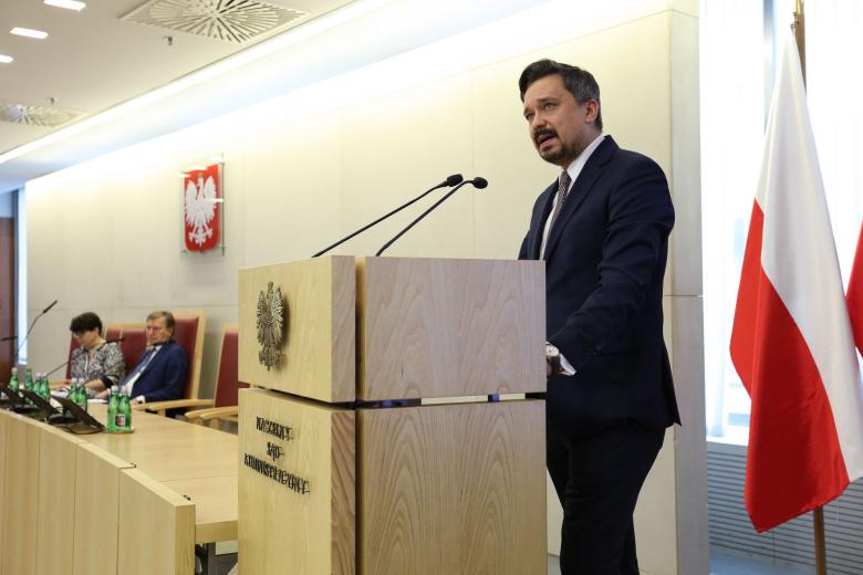 Osoba przemawia za mównicą na sali konferencyjnej, z tyłu flaga Polski