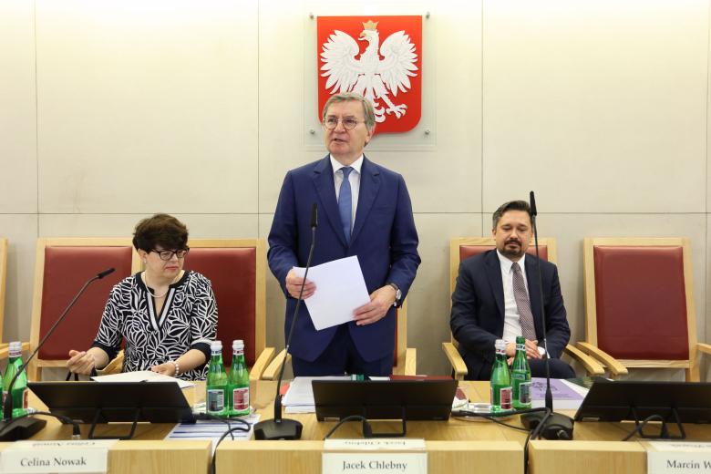 Trzy osoby za stołem prezydialnym, jedna osoba stoi i mówi do mikrofonu, na ścianie za plecami godło Polski