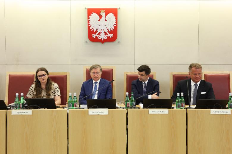 Cztery osoby siedzą za stołem prezydialnym, na ścianie za nimi godło Polski - biały orzeł w koronie na czerwonym tle